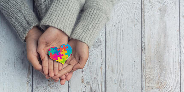 Quatro mãos juntas segurando um coração de papel com as cores da conscientização ao transtorno do espectro autista