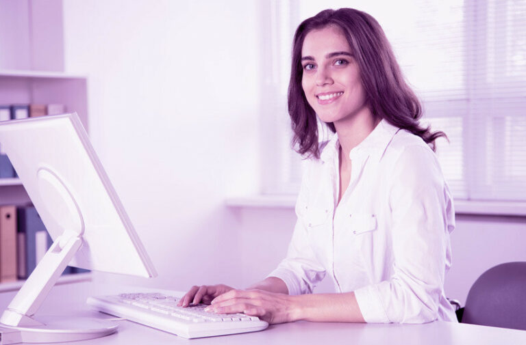 Jovem recepcionista de camisa branca sorri enquanto digita em computador na recepção da clínica, ilustrando gestão com sistema informatizado