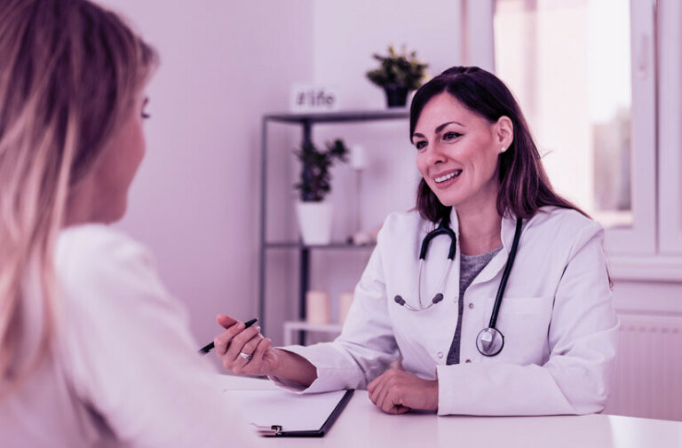 Médica sorridente de jaleco branco conversa com paciente durante consulta acolhedora, representando o atendimento adaptado às mudanças de comportamento
