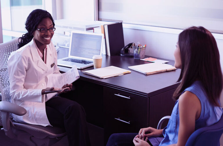 Medica sorridente de jaleco branco conversa com paciente sentada do outro lado da mesa com notebook e documentos
