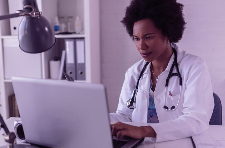 Médica de jaleco e estetoscópio concentrada digitando no notebook em consultório, representando pesquisas em saúde com uso da CID 11