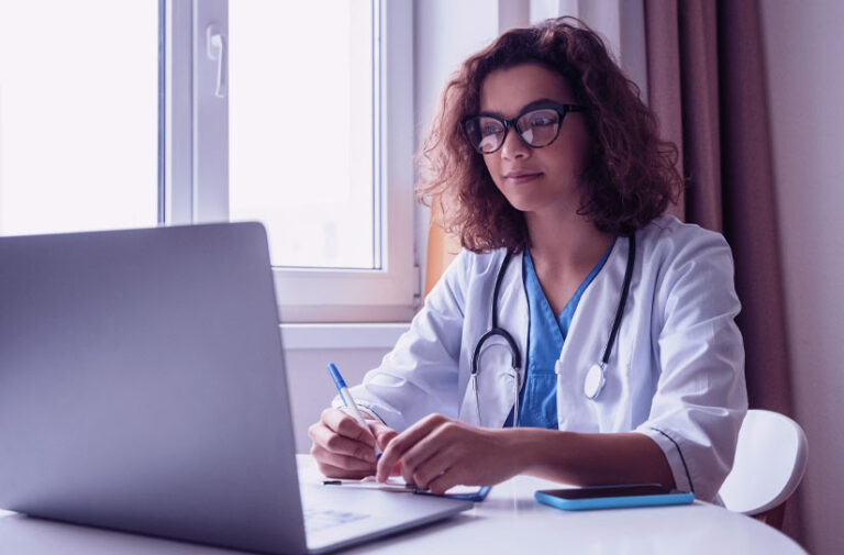 Jovem médica de óculos e jaleco fazendo anotações à mesa com notebook e celular, aplicando a classificação CID 11 no dia a dia