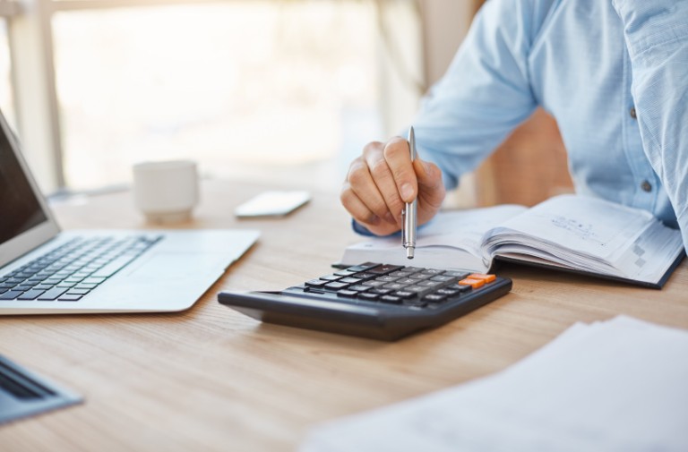 Homem de camisa azul usa calculadora e anota valores em caderno ao lado de notebook, elaborando orcamentos financeiros