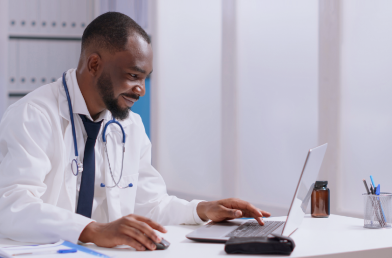 Medico negro de jaleco trabalha no notebook em consultorio, acessando documentos com o software da Amplimed