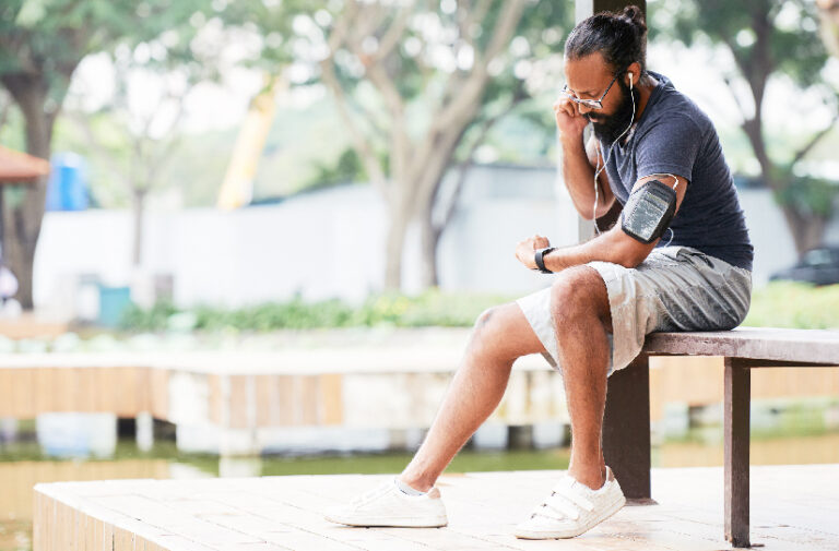 Homem sentado em banco de parque ajusta fone de ouvido usando smartwatch e braceira de celular, representando gadgets para saude