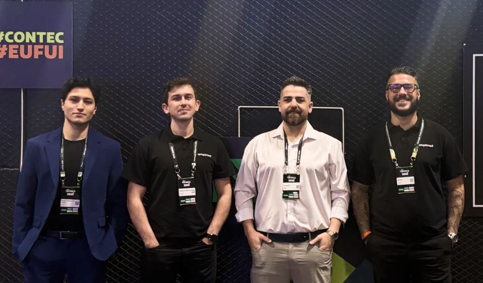 Quatro profissionais da equipe Amplimed posam juntos com crachás no estande da Convenção Técnica Unimed 2026, painel #Contec ao fundo